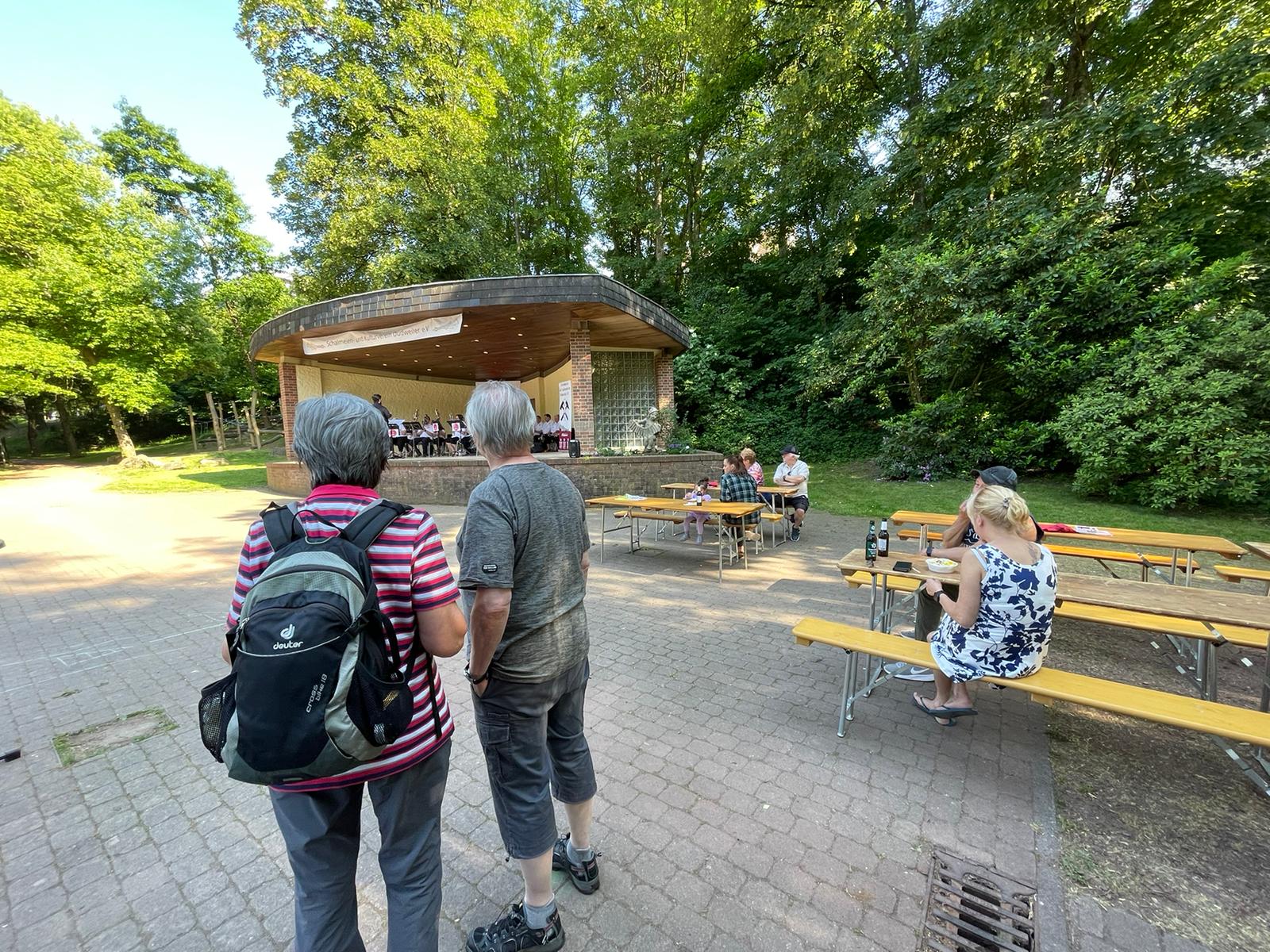 Foto im Stadtpark, man sieht den Pavillon, ein paar Bierzeltgarnituren an denen Menschen sitzen und im Vordergrund zwei Menschen von hinten, die zur Bühne schauen.
